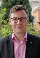 Eine Wissenschaftlerin und ein Wissenschaftler arbeiten hinter einer Glasfassade und mischen Chemikalien mit Großgeräten.